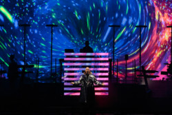 Neil Tennant of Pet Shop Boys performs in a silver metallic raincoat. Chris Lowe is seen in silhouette in front of a bright LED screen in the background at Budweiser Stage in Toronto, CANADA