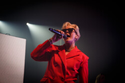 Australian-American singer Betty Who performs on stage in a red suit and sunglasses, holding a microphone