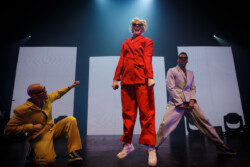 Australian-American singer Betty Who performs on stage in a red suit and sunglasses, holding a microphone, flanked by two male dancers