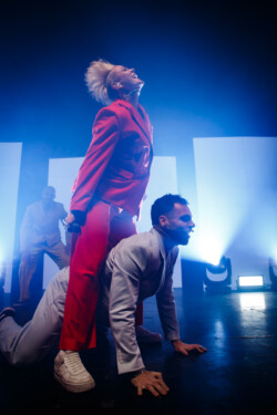Australian-American singer Betty Who performs on stage in a red suit holding a microphone straddling a dancer who is on all fours