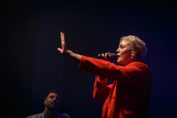 Australian-American singer Betty Who performs on stage in a red suit holding a microphone with a backup dancer behind her