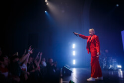 Australian-American singer Betty Who performs on stage in a red suit holding a microphone while a crowd cheers