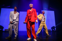 Australian-American singer Betty Who performs on stage in a red suit holding a microphone, flanked by two male dancers