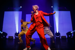 Australian-American singer Betty Who performs on stage in a red suit holding a microphone, flanked by two male dancers