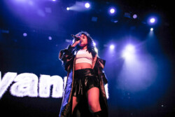 Aáyanna wearing an oversize leather jacket, white tank top and black leather skirt performs on stage holding a microphone
