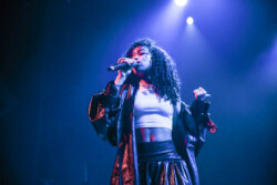 Aáyanna wearing an oversize leather jacket, white tank top and black leather skirt performs on stage holding a microphone