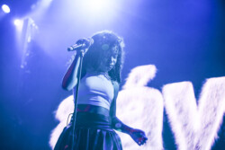 Aáyanna wearing a white tank top and black leather skirt performs on stage holding a microphone stand