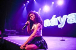 Aáyanna wearing a white tank top and black leather skirt sits at the edge of the stage holding a microphone