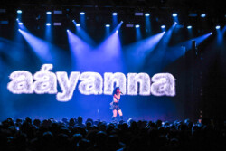 Aáyanna wearing a white tank top and black leather skirt performs on stage in front of a digital screen displaying her name in a furry font