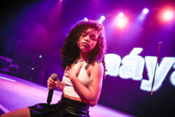 Aáyanna wearing a white tank top and black leather skirt sits at the edge of the stage holding a microphone