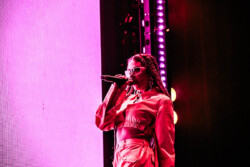 Valie of of Emotional Oranges performing in an orange jumpsuit, sunglasses and long braids