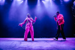 Valie of of Emotional Oranges performing in an orange jumpsuit, sunglasses and long braids