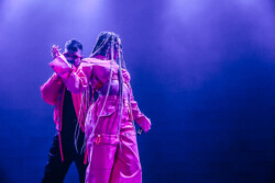 Valie of of Emotional Oranges performing in an orange jumpsuit, sunglasses and long braids
