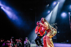 Valie of of Emotional Oranges performing in an orange jumpsuit, sunglasses and long braids
