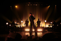 Emily King performing on stage wearing a black Bolero jacket adorned with silver buttons, a black sheer turtleneck, black pants and high heels
