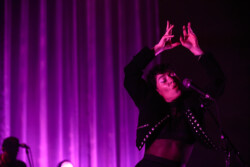 Emily King performing on stage wearing a black Bolero jacket adorned with silver buttons, a black sheer turtleneck, black pants and high heels