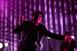 Emily King performing on stage wearing a black Bolero jacket adorned with silver buttons, a black sheer turtleneck, black pants and high heels