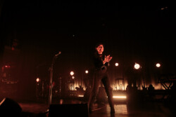 Emily King performing on stage wearing a black Bolero jacket adorned with silver buttons, a black sheer turtleneck, black pants and high heels