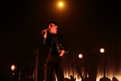 Emily King performing on stage wearing a black Bolero jacket adorned with silver buttons, a black sheer turtleneck, black pants and high heels