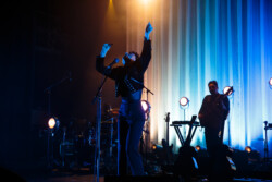 Emily King performing on stage wearing a black Bolero jacket adorned with silver buttons, a black sheer turtleneck, black pants and high heels
