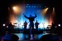 Emily King performing on stage wearing a black Bolero jacket adorned with silver buttons, a black sheer turtleneck, black pants and high heels
