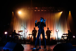 Emily King performing on stage wearing a black Bolero jacket adorned with silver buttons, a black sheer turtleneck, black pants and high heels