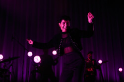 Emily King performing on stage wearing a black Bolero jacket adorned with silver buttons, a black sheer turtleneck, black pants and high heels