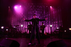 Emily King performing on stage wearing a black Bolero jacket adorned with silver buttons, a black sheer turtleneck, black pants and high heels