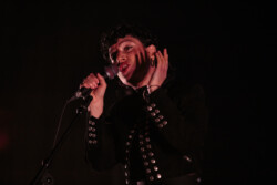 Emily King performing on stage wearing a black Bolero jacket adorned with silver buttons, a black sheer turtleneck, black pants and high heels