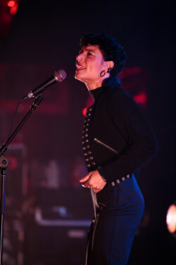 Emily King performing on stage wearing a black Bolero jacket adorned with silver buttons, a black sheer turtleneck, black pants and high heels