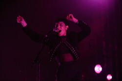 Emily King performing on stage wearing a black Bolero jacket adorned with silver buttons, a black sheer turtleneck, black pants and high heels