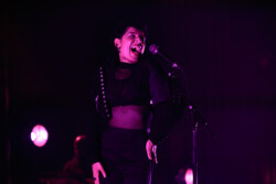 Emily King performing on stage wearing a black Bolero jacket adorned with silver buttons, a black sheer turtleneck, black pants and high heels