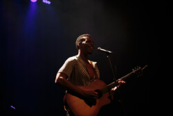 Joseph Solomon sings into a microphone on stage playing an acoustic guitar, wearing a black and white patterned shirt