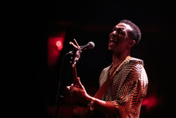 Joseph Solomon sings into a microphone on stage playing an acoustic guitar, wearing a black and white patterned shirt