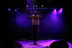 Joseph Solomon sings into a microphone on stage playing an acoustic guitar, wearing a black and white patterned shirt