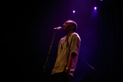Joseph Solomon sings into a microphone on stage playing an acoustic guitar, wearing a black and white patterned shirt