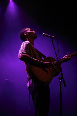 Joseph Solomon sings into a microphone on stage playing an acoustic guitar, wearing a black and white patterned shirt